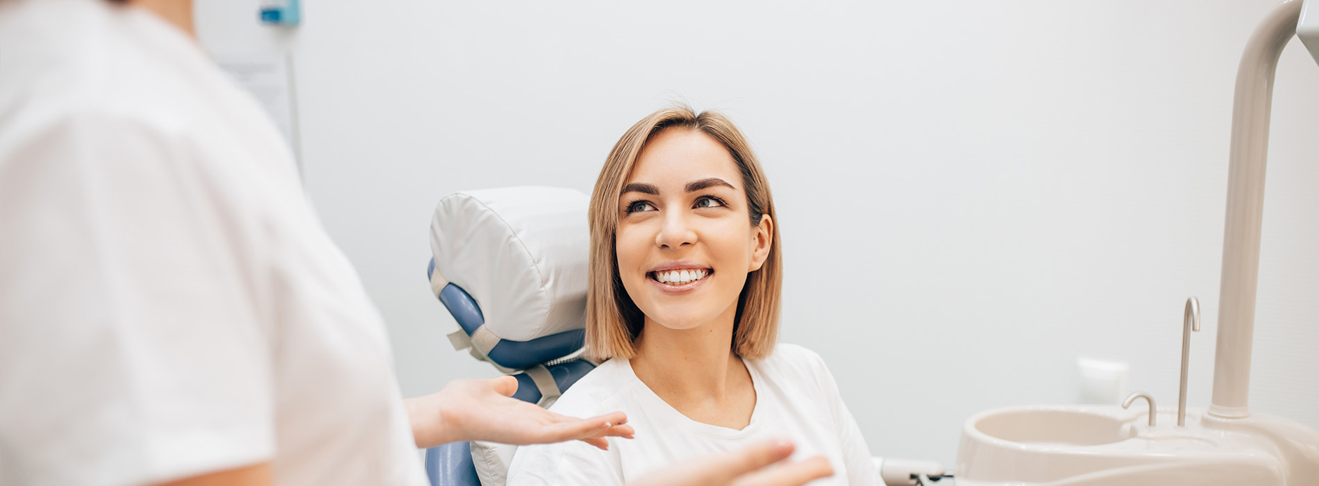 The image shows a woman sitting in a dental chair with a smile, receiving dental care from a professional wearing a white coat, who appears to be conducting an examination or procedure.