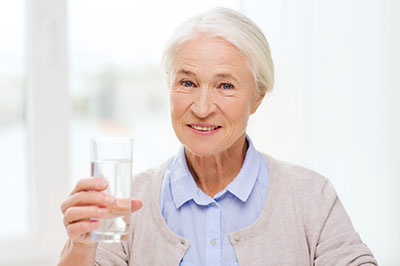 The image shows an elderly woman holding a glass of water with both hands, smiling at the camera, and standing indoors with a window behind her.