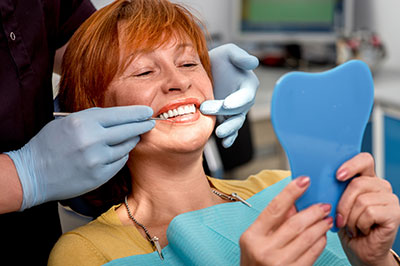 The image shows a woman sitting in a dental chair with her teeth being examined by a dentist who is holding up a tooth model, while she holds a blue mouthguard.