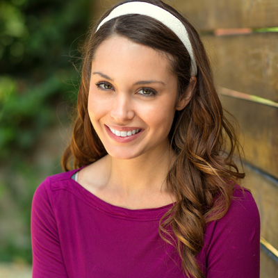 Woman with long brown hair wearing a purple top and a white headband, smiling at the camera.