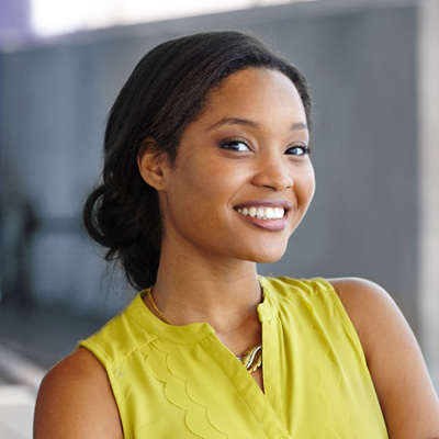 The image shows a woman with a bright smile, wearing a yellow top, posing against a concrete wall.
