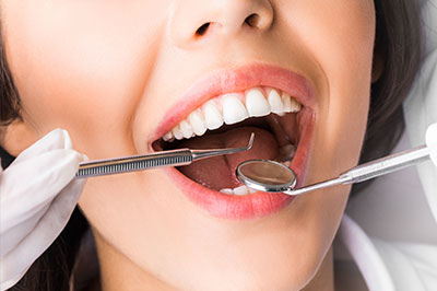 A woman s mouth with teeth being worked on by dental instruments, held open by her own hand, while she wears a surgical mask.