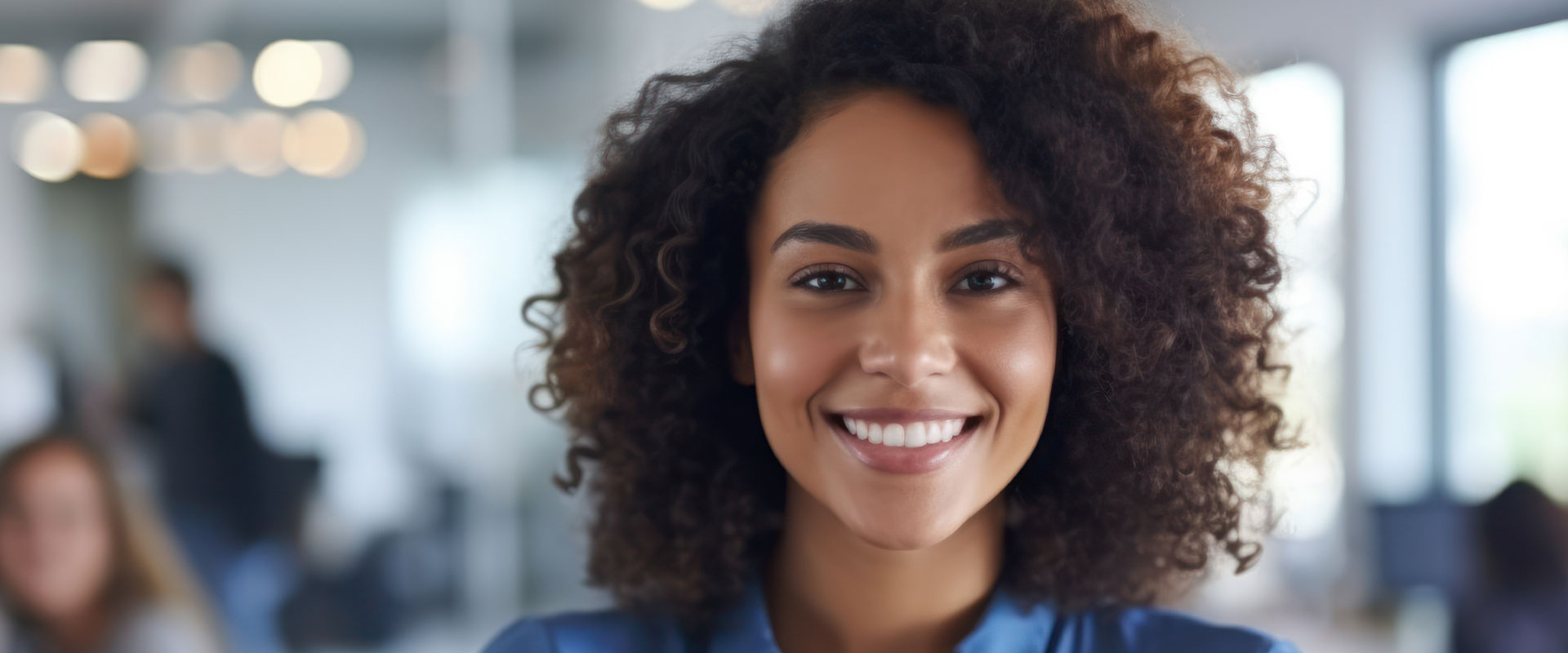 The image shows a smiling woman with curly hair, wearing a dark top, standing in an indoor setting with people behind her. She has a friendly expression and appears to be engaged in a positive interaction.