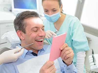 The image depicts a man sitting in a dental chair with his mouth open, holding up a pink card, while a dentist and dental hygienist look on with smiles.
