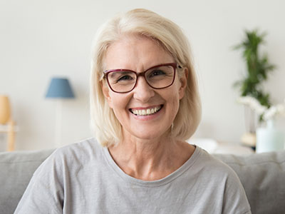 The image shows a woman with short blonde hair, wearing glasses and a light-colored top, smiling at the camera with a warm expression. She appears to be indoors, possibly in a home setting, with a white wall and a plant visible behind her.