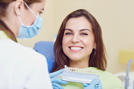 A woman seated in a dental chair receiving dental care from a professional wearing a surgical mask and holding a dental tool, with both smiling at the camera.