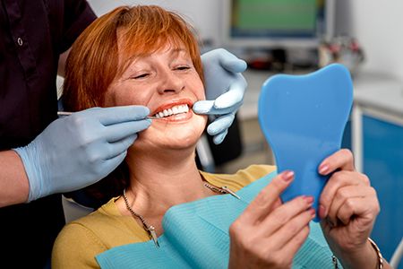 The image shows a woman seated at a dental chair, holding up a blue toothbrush, with her mouth open wide, receiving dental care from a dental professional who appears to be adjusting her teeth.